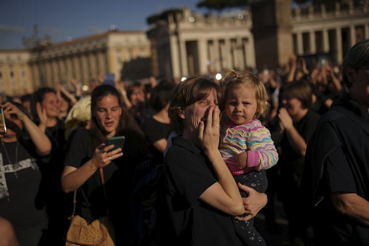 A woman puts her hand over her mouth as she holds a young girl amid a crowd of people outside.