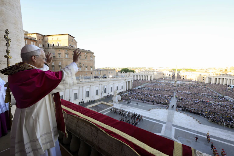 A man in a white skullcap, short red cape and white robe waves to a vast crowd outside.