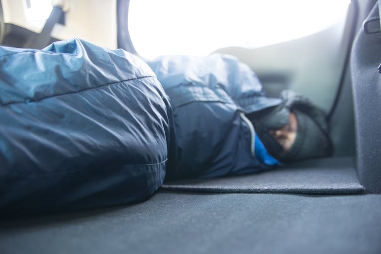 A man sleeps in a sleeping bag inside a car.