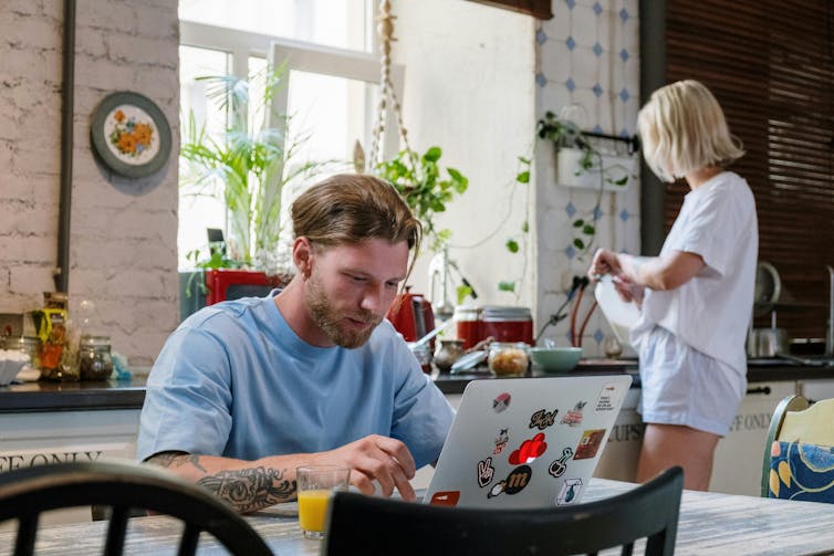 A young man on a laptop at home, with a woman in the background in the kitchen.