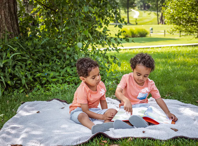 Twin boys read a book on a picnic rug.
