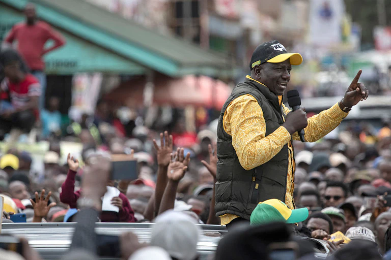 A man standing from the roof of a car speaking into a microphone while surrounded by people.