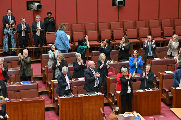 A crowd of senators stand in the chamber and applaud