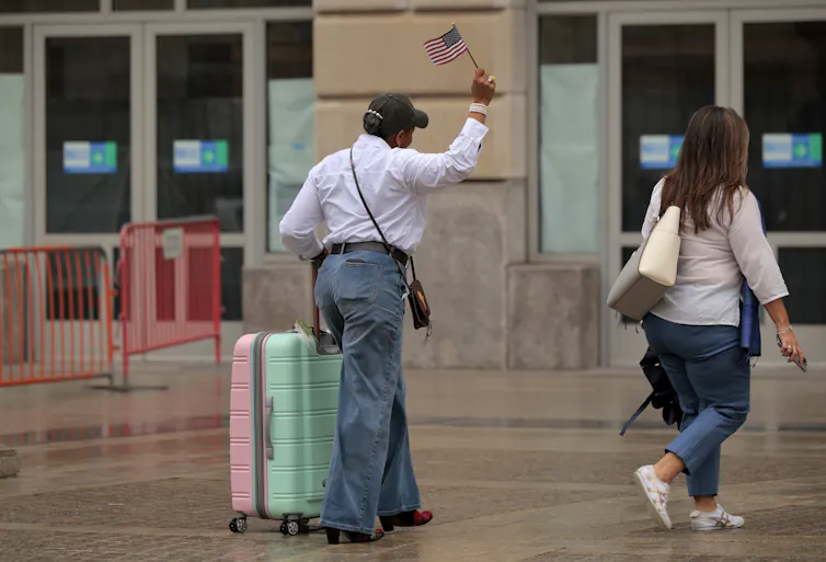 Two people are seen walking from behind. One of them rolls a suitcase and waves a small American flag.