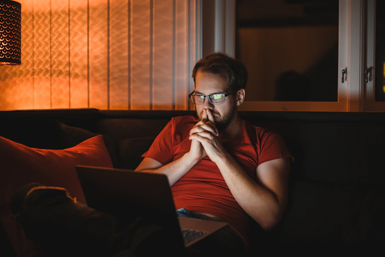 Um jovem com barba e óculos sentado em um sofá, olhando atentamente para o computador em seu colo. Suas mãos estão cruzadas em frente ao rosto.