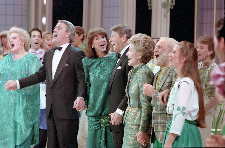 Women in green and men in black tuxedos stand on stage, their mouths wide open and singing.