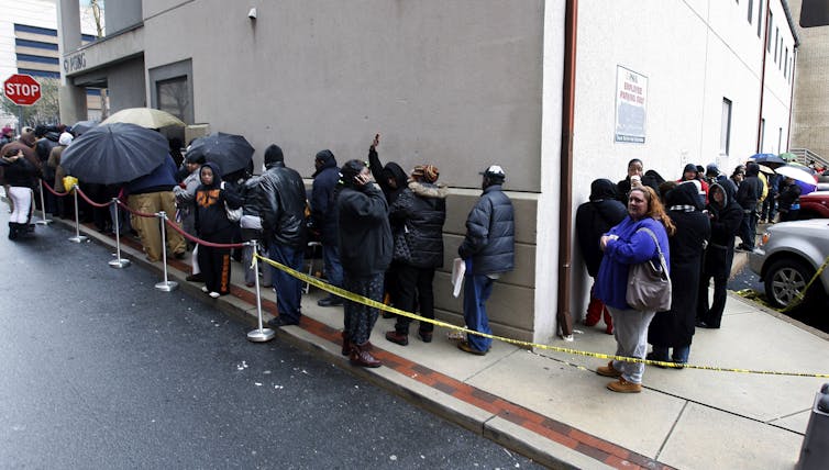 People wait in a line going around a building. Some have umbrellas.