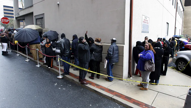 People wait in a line going around a building. Some have umbrellas.