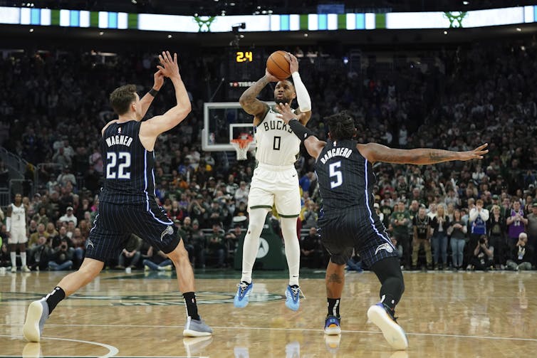 A man in a white basketball uniform throwing the ball between two men in dark blue uniforms