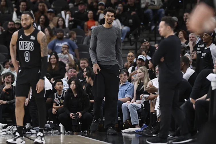 A tall man in a grey shirt standing at the side of a basketball court