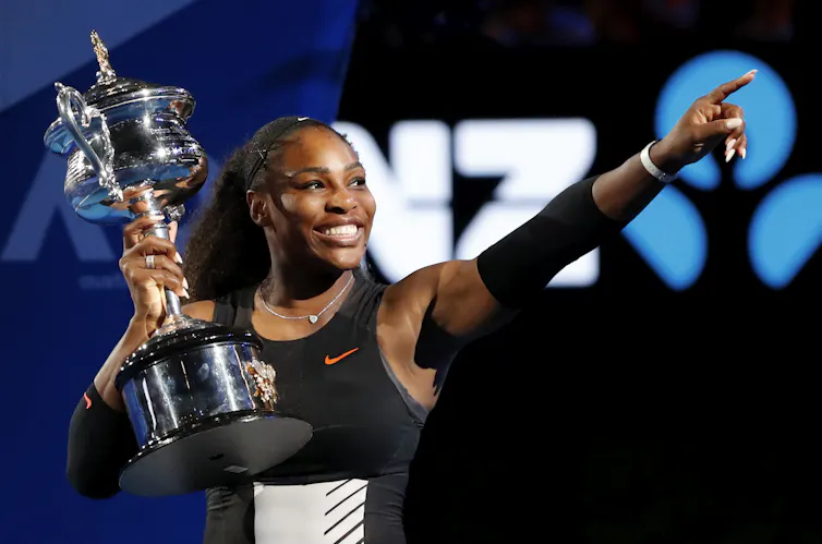 Serena Williams holding a large trophy, smiling and pointing upwards