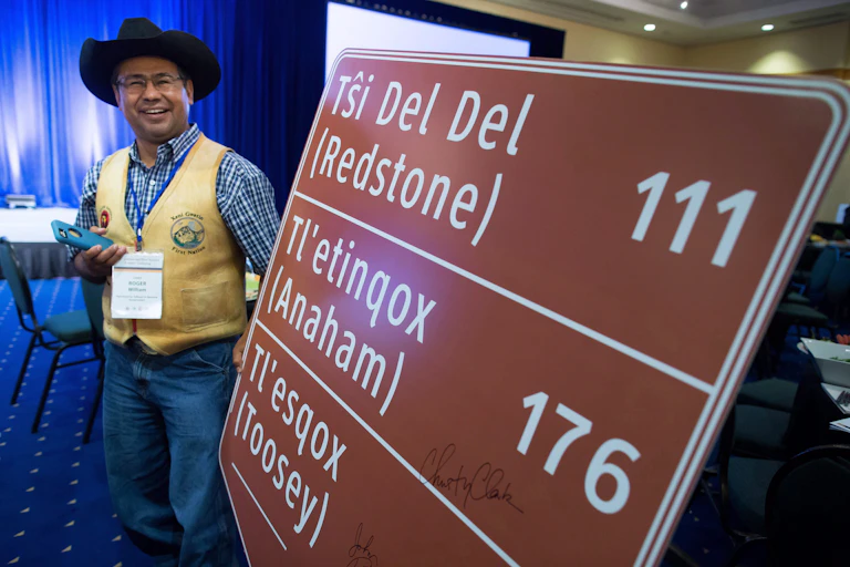 A smiling Indigenous man wearing a cowboy hat stands next to a sign featuring the names of town in the Tsilhqot’in language.