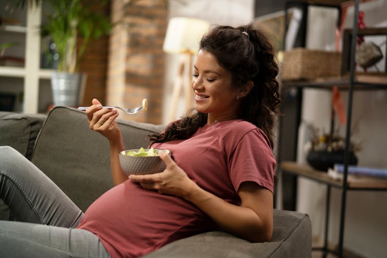 A pregnant woman sitting on a sofa eating a healthy snack.