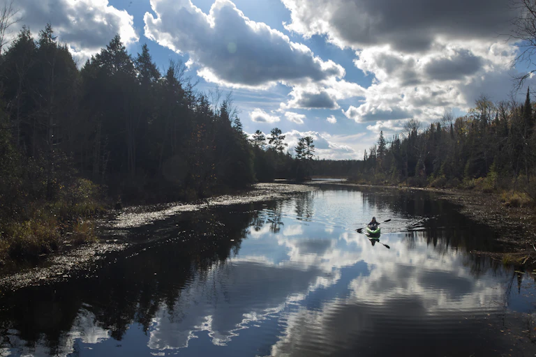 A woman kayaks on a calm body of water with dense forest on either side and clouds illuminated by the sun in front of her.