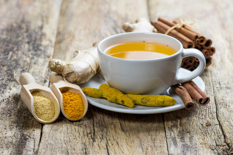 Chá herbal de gengibre e cúrcuma em xícara branca com raiz de gengibre, cúrcuma seca, paus de canela e seu pó.