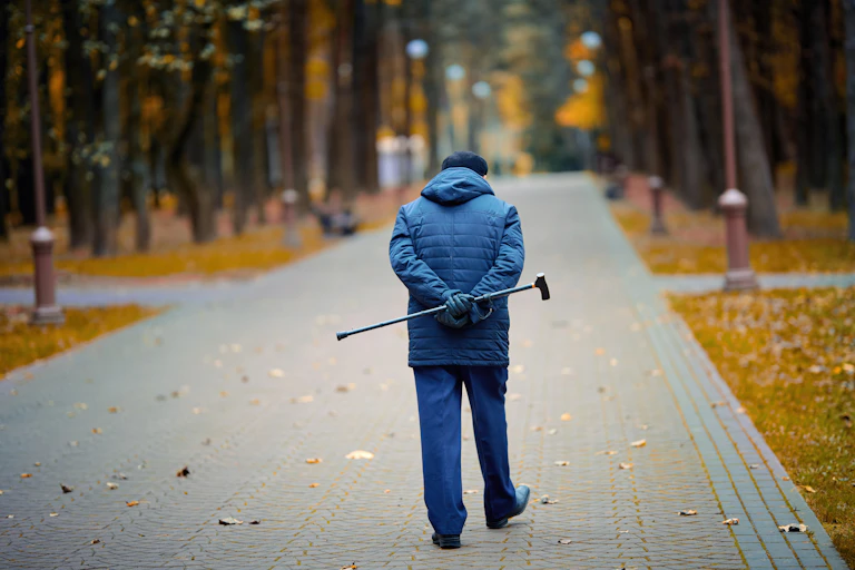 Elderly man walking down a path in autumn, holding a walking stick behind his back.