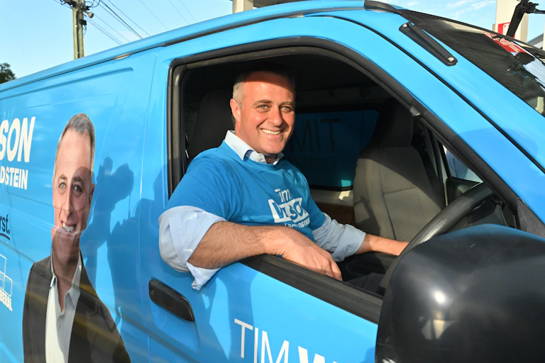 Tim Wilson, dressed in a blue Liberal Party shirt, sitting inside a blue Liberal Party van.