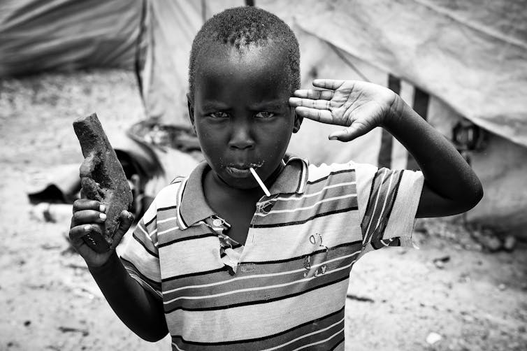 A young boy gives a military salute and holds up a pistol made of mud.