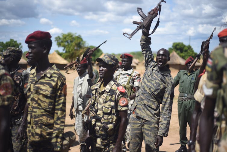 A young uniformed soldier amid a troop lifts his rifle above his head and shouts.