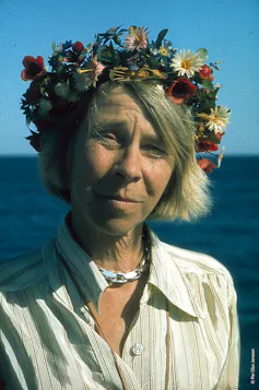 An older Finnish woman wearing a white blouse with a flower garland crown on her head.