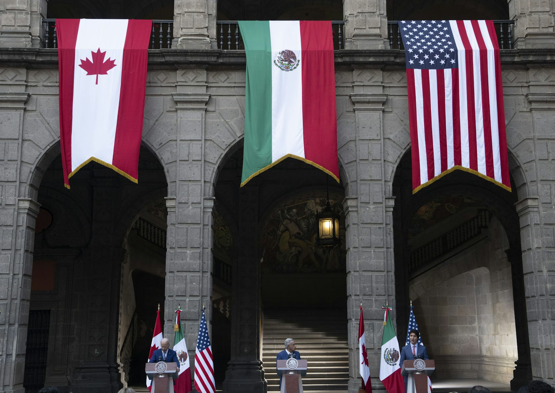 Banderas canadienses, mexicanas y estadounidenses que cuelgan de la parte superior del edificio. A continuación, el hombre se encuentra debajo de cada bandera en el podio.