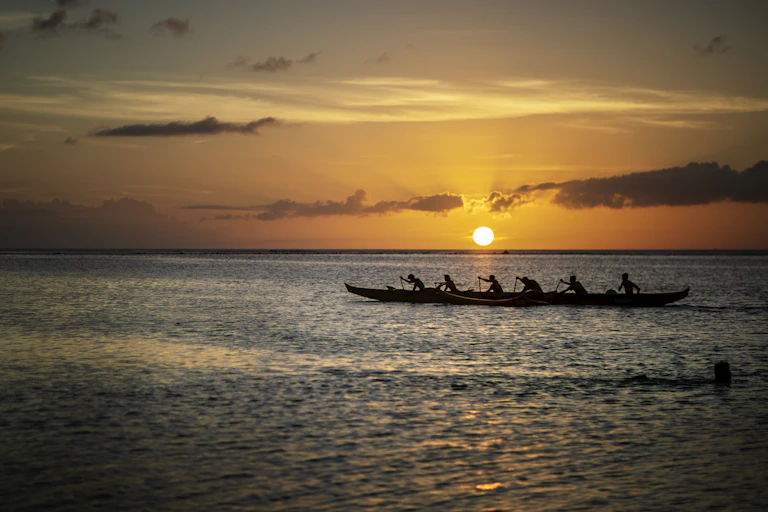 silhouette of long canoe with six people in it, against setting sun in distance