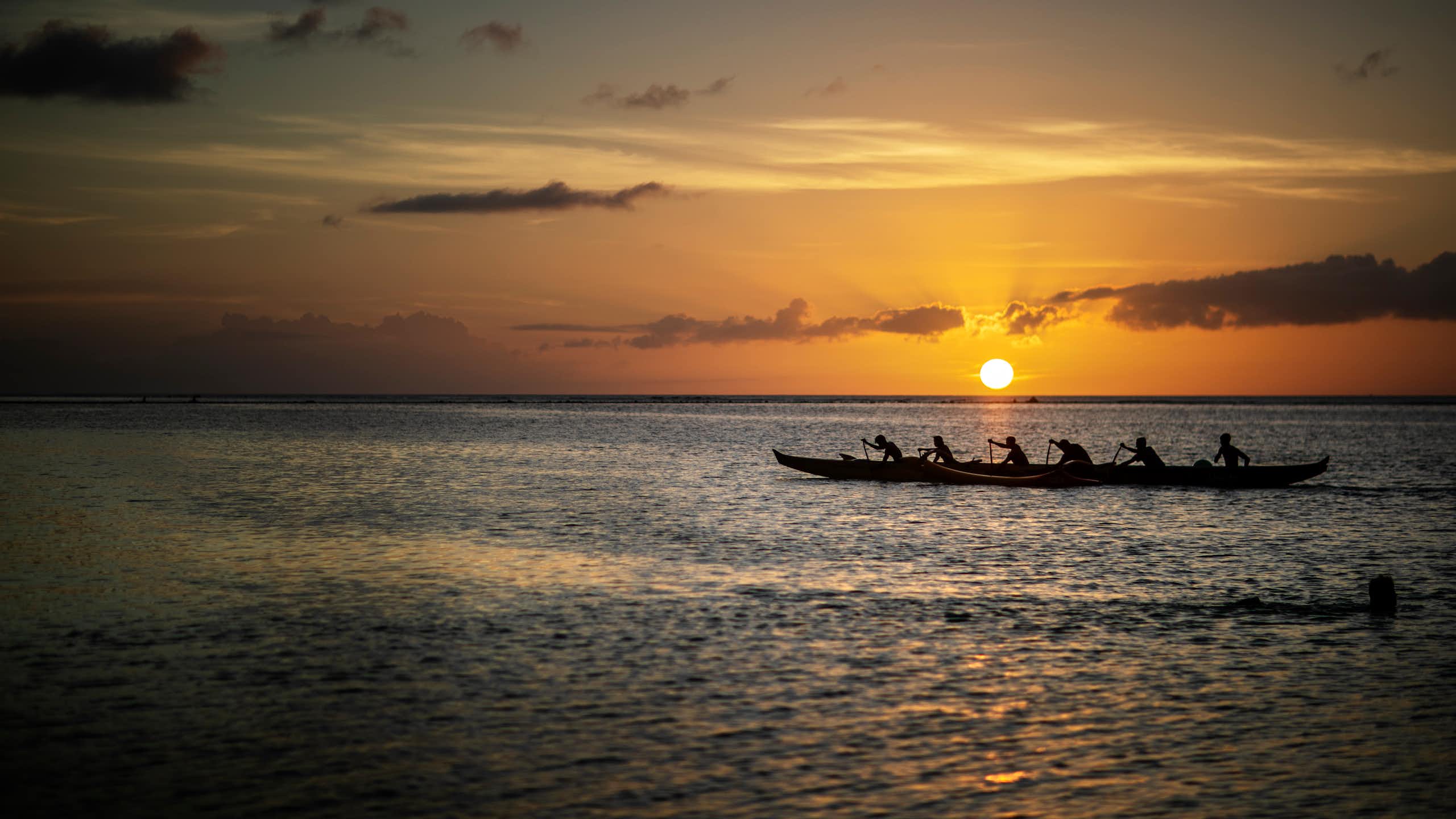 silhouette of long canoe with six people in it, against setting sun in distance