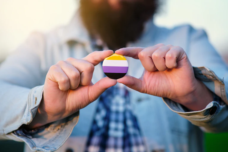 A person with a beard and button-up shirt, whose face is outside the photo frame, holds up a pin with stripes of yellow, white, purple and black.