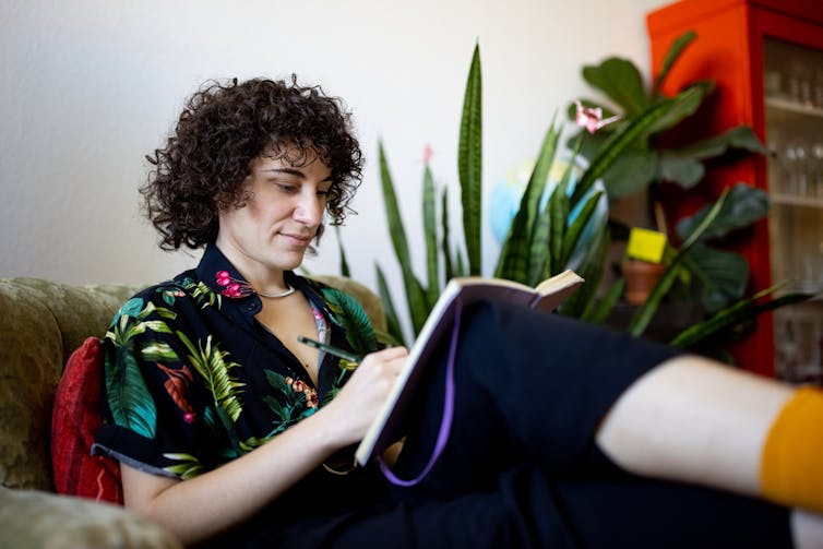 A person with curly hair, who is wearing a black and floral shirt, sits while writing in a journal.