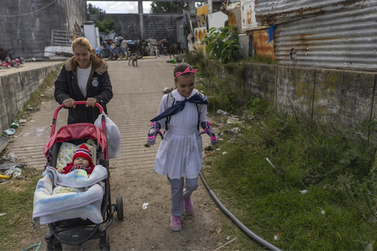 a girl with brightly colored prosthetic hands walks beside a woman pushing a baby in a stroller