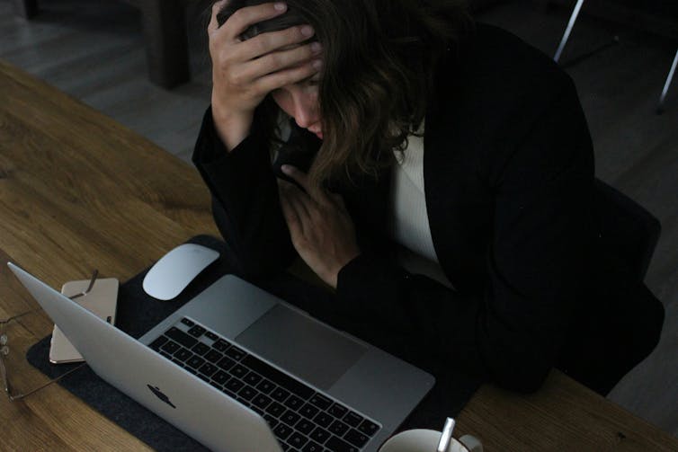 A woman in a suit sits at a table in front of an open laptop with her head in her hands