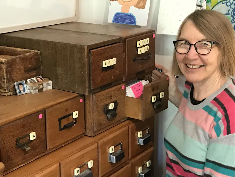 Smiling woman with glasses and a striped jumper, with a wooden filing cabinet with index cards.
