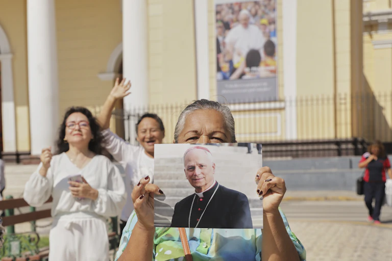 Someone wearing a bright green shirt holds up a large photograph of a man in black robes outside a cream-colored building.