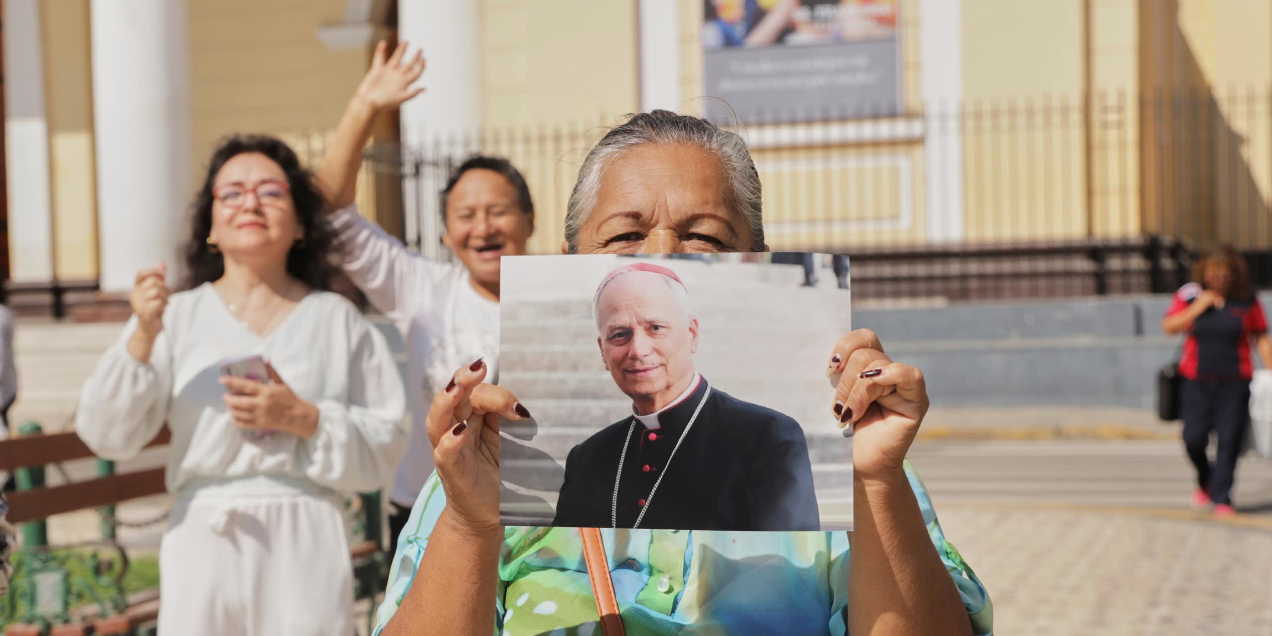 Someone wearing a bright green shirt holds up a large photograph of a man in black robes outside a cream-colored building.