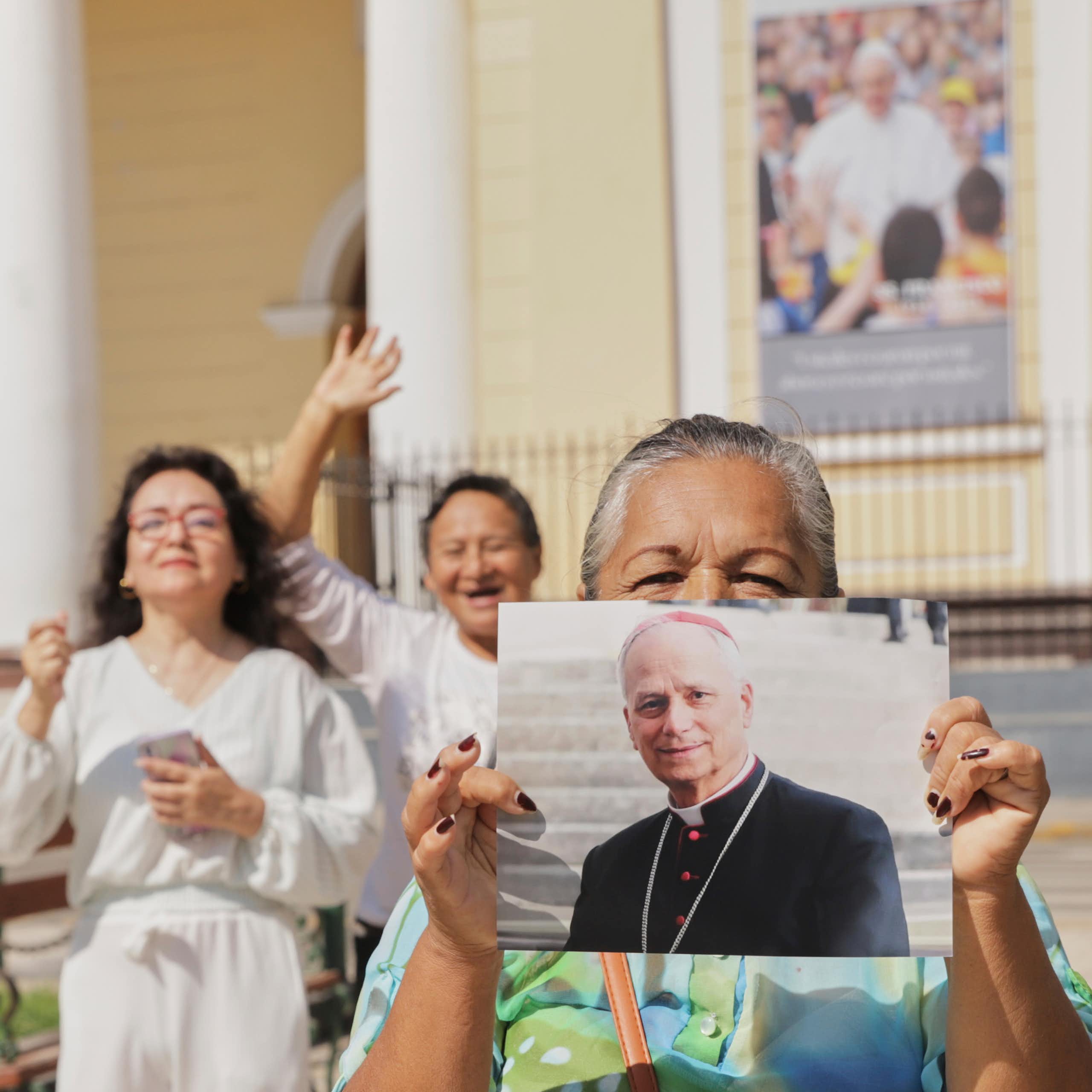 Someone wearing a bright green shirt holds up a large photograph of a man in black robes outside a cream-colored building.