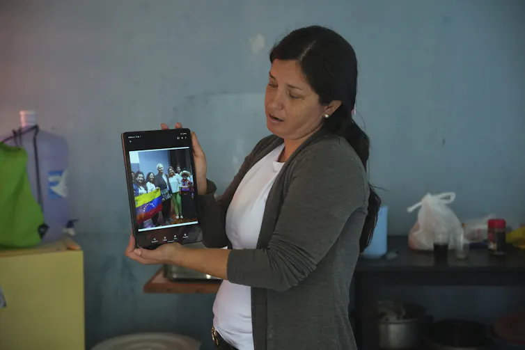 A woman in a white shirt and gray sweater holds up an electronic tablet displaying a picture.