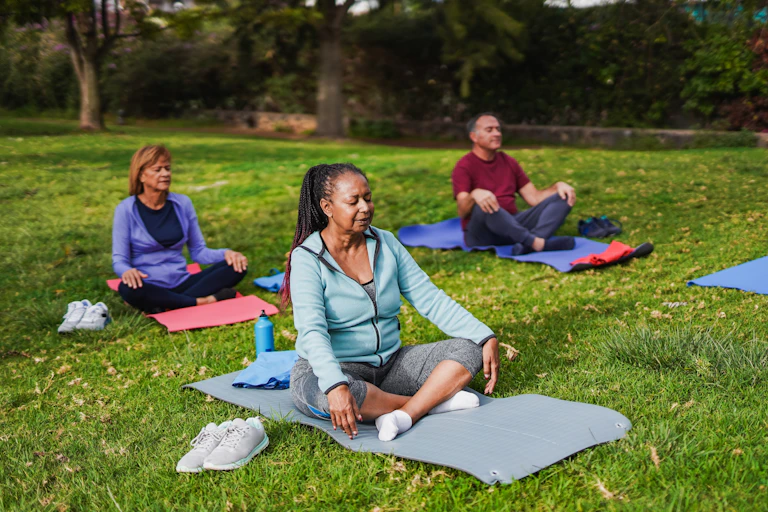 Three middle-aged or older adults do yoga in the park while sitting on their yoga mats.