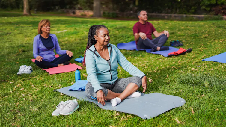 Three middle-aged or older adults do yoga in the park while sitting on their yoga mats.