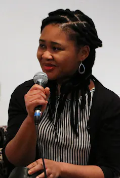 A young African woman with dreadlocks speaks into a microphone.