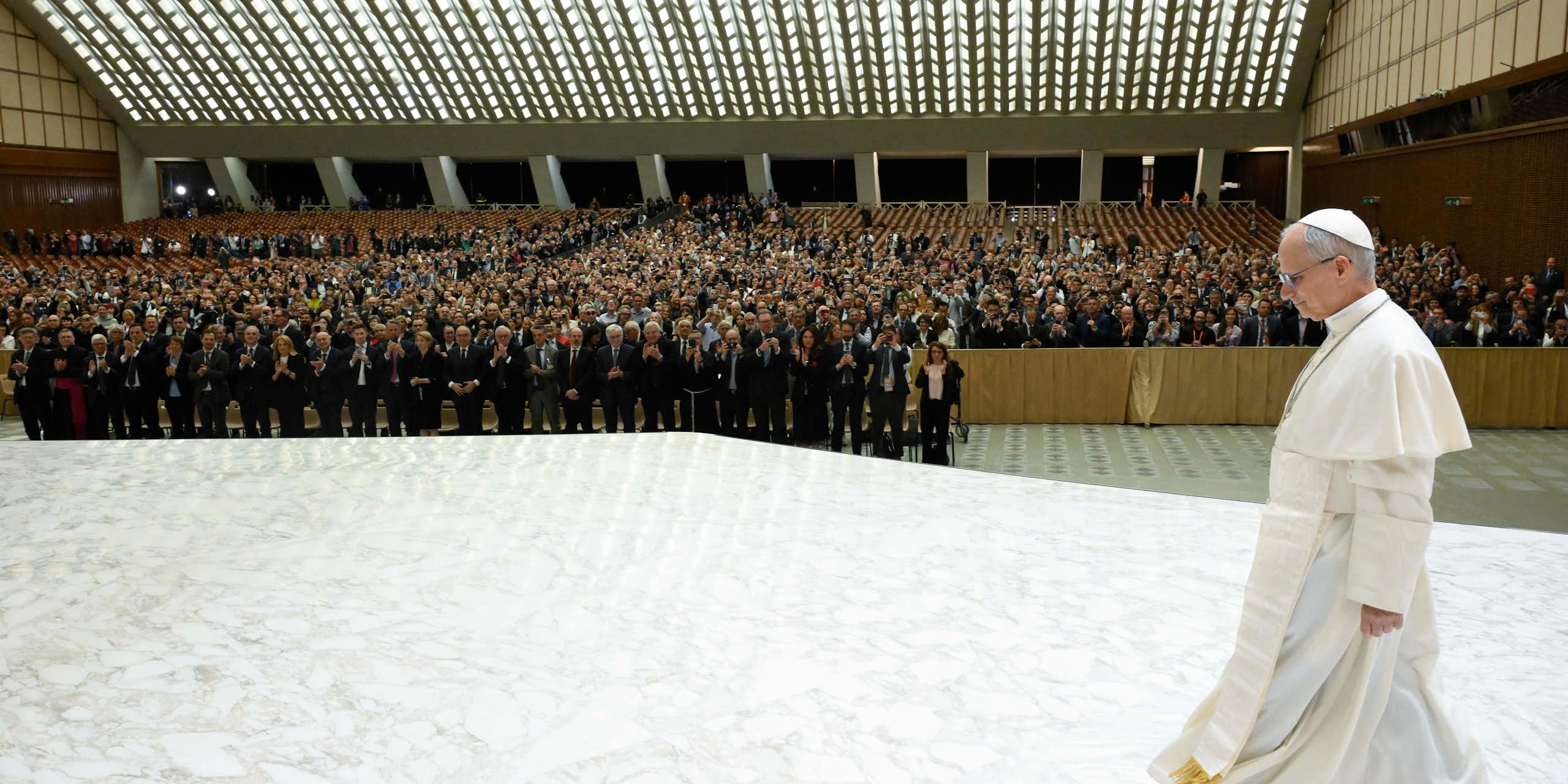 Man in white robe strides across a stage before a huge crowd of people.