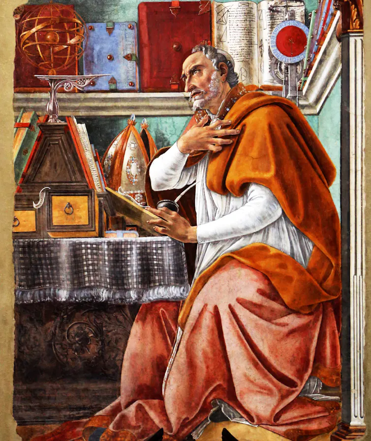 Un homme vêtu de blanc et d’ocre, assis à une table, tient un livre ouvert et regarde devant lui