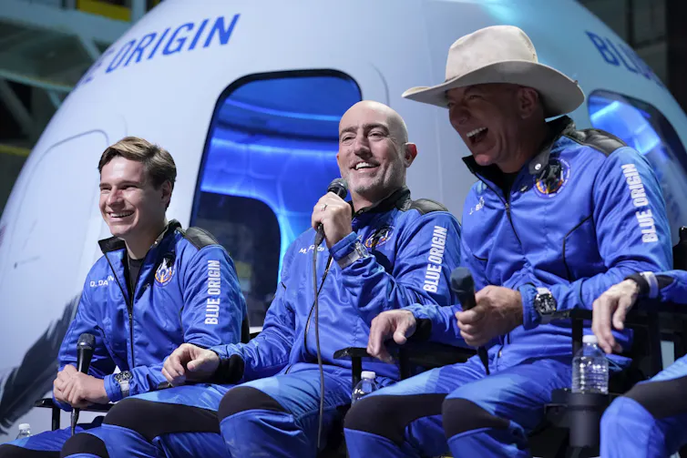 Three men -- including Jeff Bezos in a cowboy hat -- wear blue jumpsuits and sit in front of a model crew capsule labeled 'Blue Origin.'