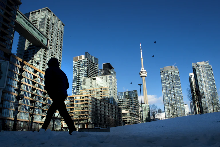 A person in a coat walks through snow as the Toronto skyline is seen in the background