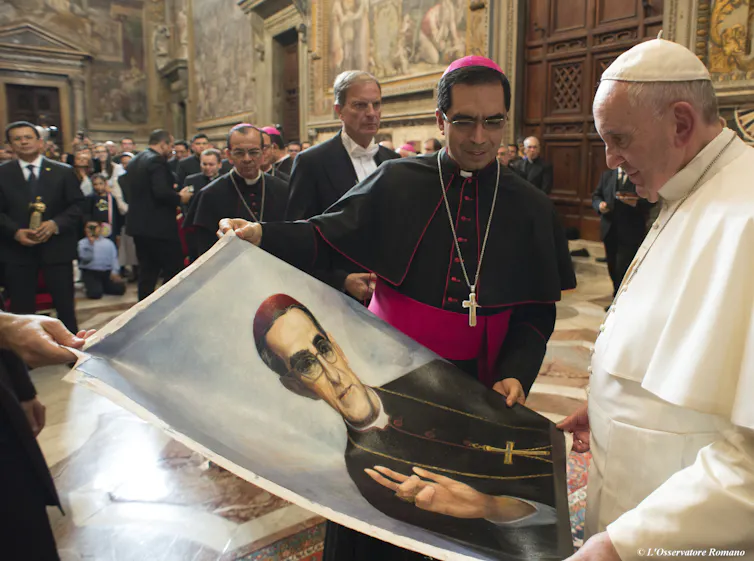 A man in a white robe, cape and skullcap looks at a poster of a priest in a black robe, held by two other men.