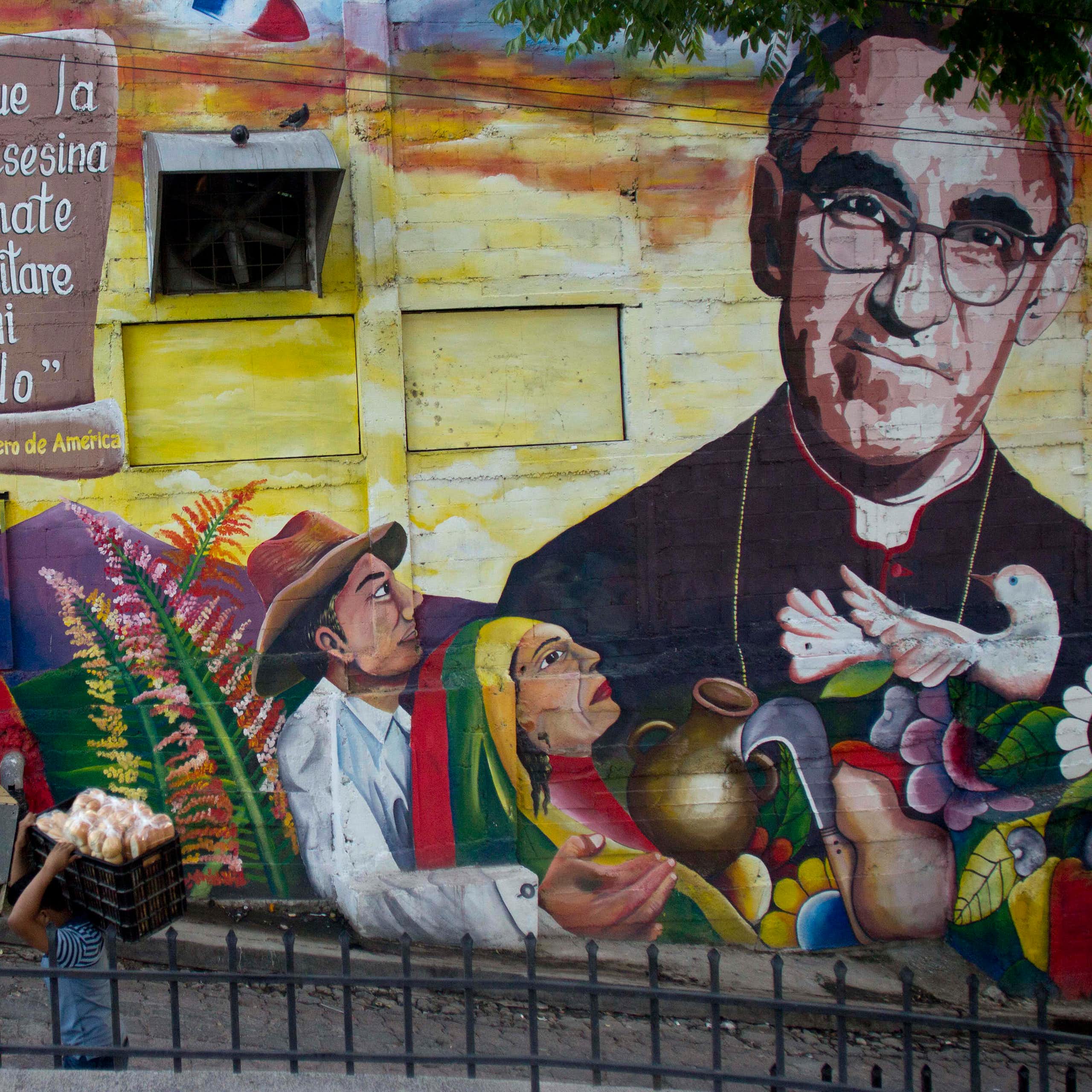 A man walks past an outdoor mural showing several figures, including one in a black priest's robe.