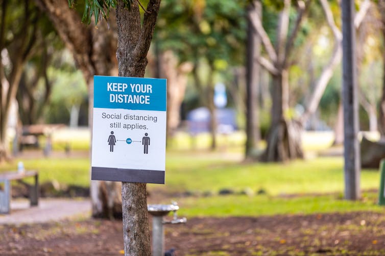 A COVID-19 sign with the words 'Keep Your Distance' nailed to a tree in a park.