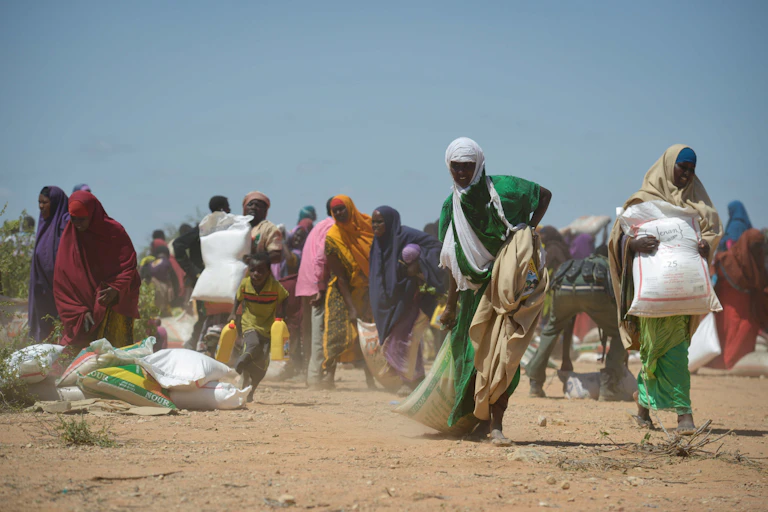 A woman dragging a large sack as other women around her walk away with white sacks picked up from the dusty ground.