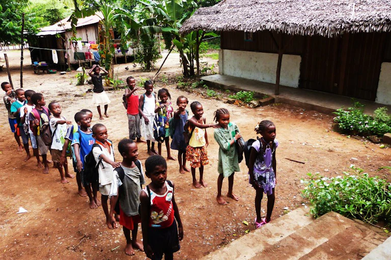 Two lines of young children standing in front of the steps up to a building. Small thatched wooden buildings and palm trees in the background.