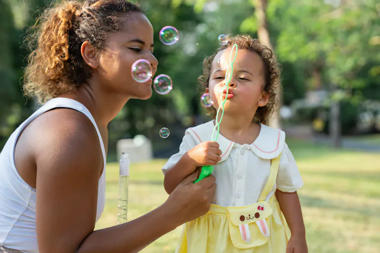 Young woman and a child blow bubbles together in a park.