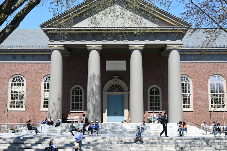 People sit on stairs and on chairs in front of a building on a college campus.